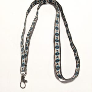 May include: A fabric lanyard featuring a repeating geometric pattern in shades of blue, brown, and cream. The strap is attached to a silver-toned metal swivel lobster clasp at the bottom, designed for holding items securely with a durable woven texture.