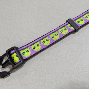 May include: A purple and black pet collar with a repeating pattern of green Frankenstein monster faces. The collar has a black plastic buckle and is designed for a dog or cat.