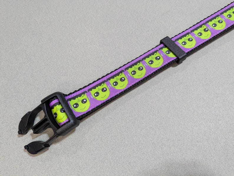 May include: A purple and black pet collar with a repeating pattern of green Frankenstein monster faces. The collar has a black plastic buckle and is designed for a dog or cat.