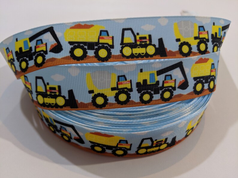 May include: A blue and brown ribbon with a repeating pattern of yellow construction vehicles. The vehicles include a dump truck, an excavator, and a bulldozer.