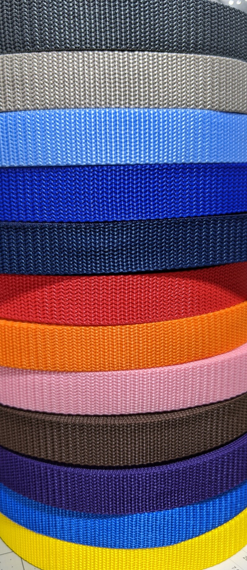 May include: A stack of 13 different colored webbing straps. The straps are all the same width and are arranged in a stack from top to bottom. The colors are black, gray, light blue, blue, navy blue, red, orange, pink, brown, purple, royal blue, and yellow.