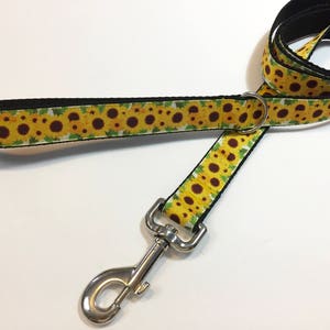 May include: A yellow and green dog leash with a sunflower pattern. The leash has a silver clip and a black strap.