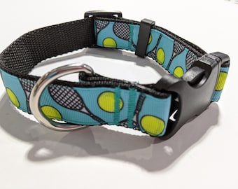 May include: A blue dog collar with a tennis racket and ball pattern. The collar has a black buckle and a silver D-ring.