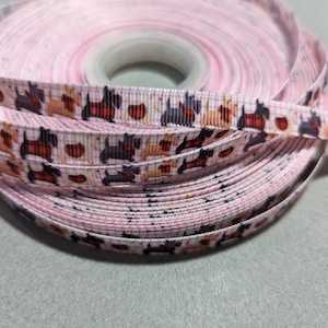 Può includere: Una bobina di nastro grosgrain rosa chiaro con un motivo ripetuto di piccoli cani terrier scozzesi neri e marrone chiaro che indossano cappotti a quadri rossi, intervallati da piccole forme circolari rosse, avvolta attorno a un nucleo centrale.