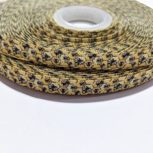 May include: A roll of decorative ribbon with a repeating pattern of yellow honeycombs and black and white bees. The ribbon is narrow and tightly wound on a white spool. The ribbon is suitable for crafts.
