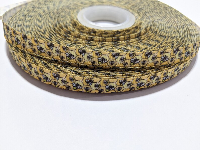 May include: A roll of decorative ribbon with a repeating pattern of yellow honeycombs and black and white bees. The ribbon is narrow and tightly wound on a white spool. The ribbon is suitable for crafts.