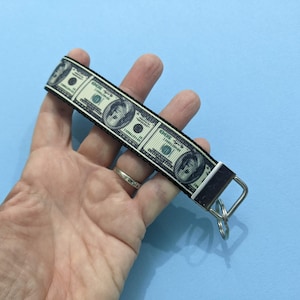 May include: A money-themed keychain featuring a repeating pattern of $100 bills. The keychain has a black border and a silver-colored metal clasp and key ring. The background is a solid light blue.