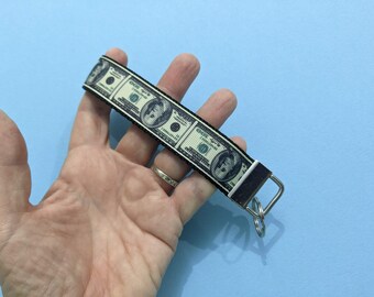 May include: A money-themed keychain with a repeating pattern of £100 bills. The keychain has a black border and a silver-coloured metal clasp and key ring. The background is a solid light blue.