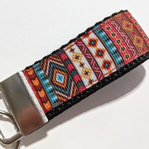 May include: A colorful key chain with a geometric pattern in shades of orange, red, blue, brown, and white. The key chain has a silver metal key ring and a black strap.