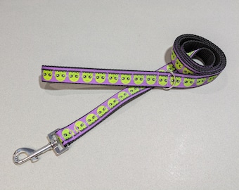 May include: A purple and black dog leash featuring a repeating pattern of green alien faces. The leash has a silver metal clasp and a matching D-ring. The leash is rolled up in a spiral.