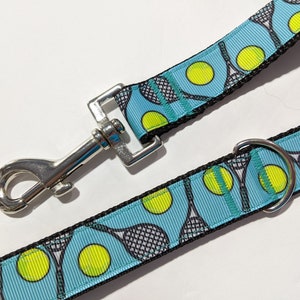 May include: A blue and black dog leash with a tennis ball and racket pattern. The leash has a silver clip and a silver ring.