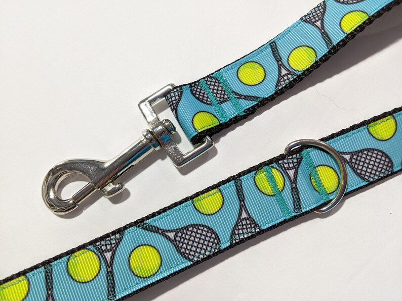 May include: A blue and black dog leash with a tennis ball and racket pattern. The leash has a silver clip and a silver ring.