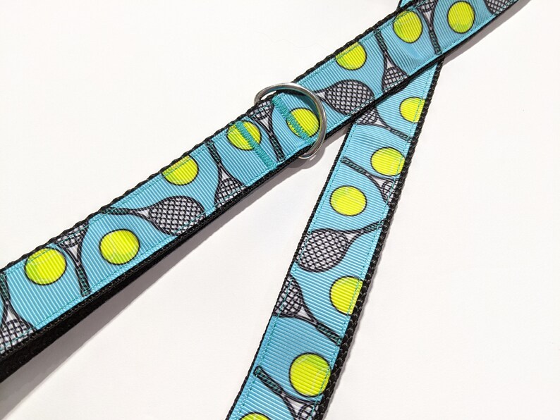 May include: A blue dog leash with a tennis racket and ball pattern. The leash has a silver metal ring.