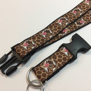 Può includere: Un cordino con stampa giraffa marrone e nera caratterizzato da un motivo ripetuto di facce di giraffa in stile cartoon con fiocchi rosa. Include una fibbia a sgancio laterale in plastica nera e un anello diviso in metallo argentato.