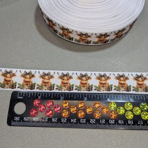 May include: White ribbon with a repeating pattern of brown and white Highland cows wearing bows. The ribbon is 1 inch wide and is shown next to a ruler.
