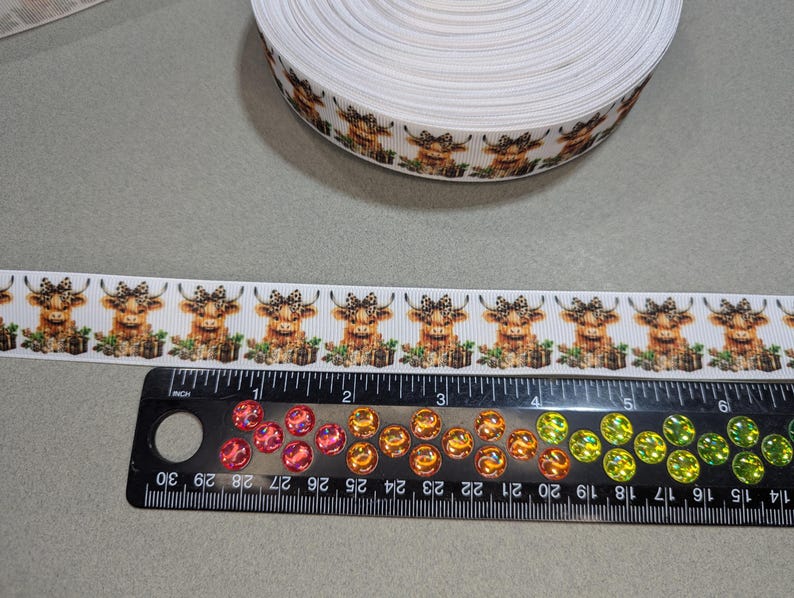 May include: White ribbon with a repeating pattern of brown and white Highland cows wearing bows. The ribbon is 1 inch wide and is shown next to a ruler.