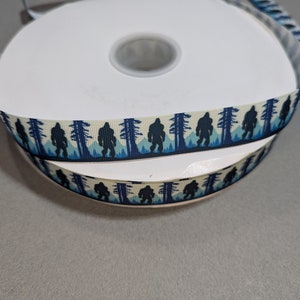 May include: A roll of white and blue ribbon with a repeating pattern of a Bigfoot silhouette walking through a forest. The ribbon is on a white spool.