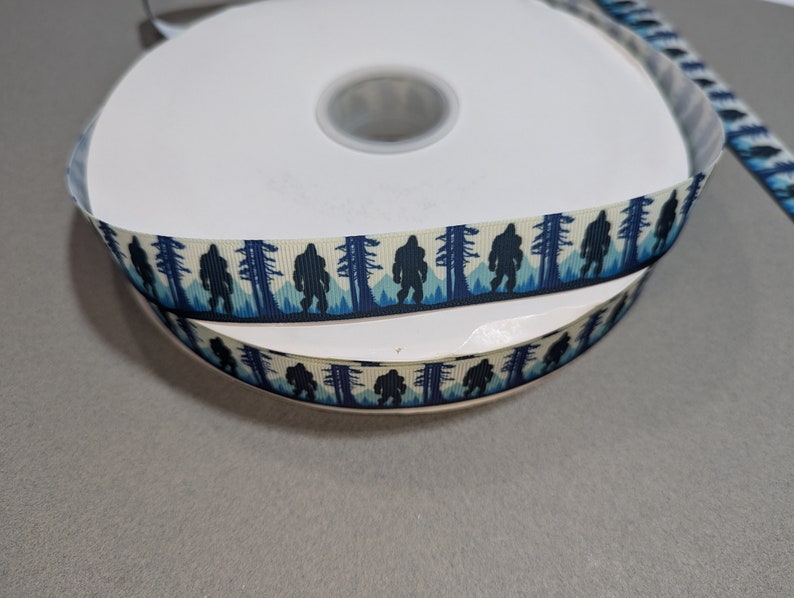 May include: A roll of white and blue ribbon with a repeating pattern of a Bigfoot silhouette walking through a forest. The ribbon is on a white spool.