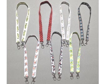 May include: A collection of seven fabric lanyards featuring various patterns including fish, red and black plaid, white with fruit prints, and geometric designs. Each lanyard is equipped with a metal swivel lobster clasp at the bottom end.