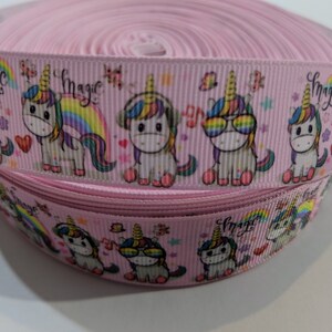 May include: A spool of light pink grosgrain ribbon featuring a repeating pattern of cartoon unicorns with colourful manes, rainbows, hearts, and the word magic printed in black script. The unicorns are depicted wearing headphones or sunglasses.