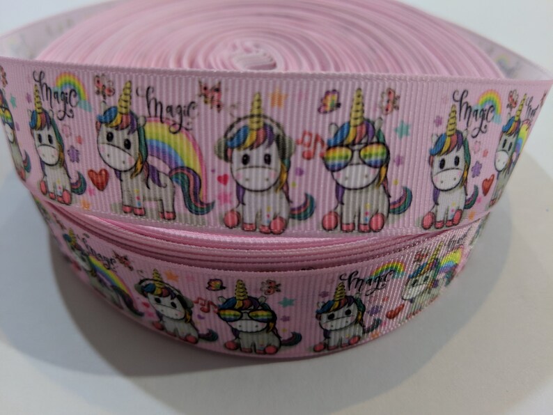 May include: A spool of light pink grosgrain ribbon featuring a repeating pattern of cartoon unicorns with colourful manes, rainbows, hearts, and the word magic printed in black script. The unicorns are depicted wearing headphones or sunglasses.