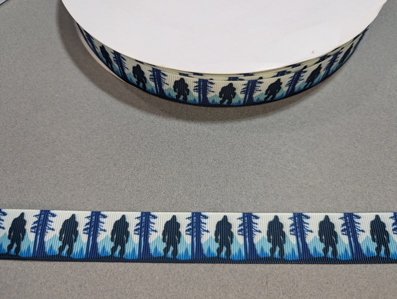 May include: A white ribbon with a repeating pattern of blue silhouette Bigfoot figures walking through a blue and white forest.