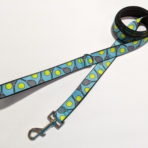 May include: A blue dog leash with a black border and a pattern of tennis rackets and tennis balls. The leash has a silver clip.