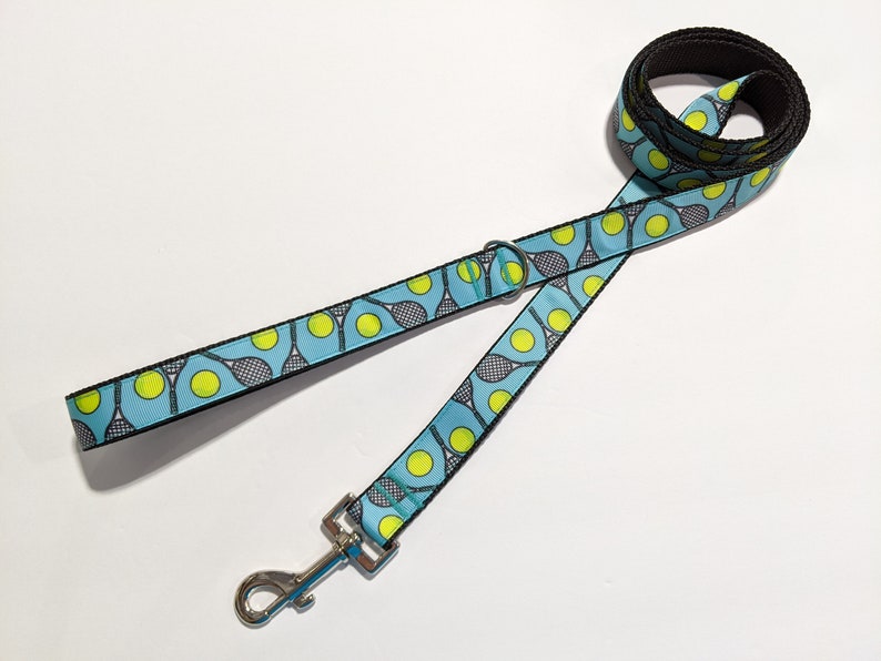 May include: A blue dog leash with a black border and a pattern of tennis rackets and tennis balls. The leash has a silver clip.