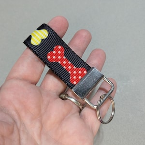 May include: Black fabric keychain with a red bone design with white polka dots and yellow paw prints. The keychain has a silver metal clasp and key ring. The design is likely for a pet owner.