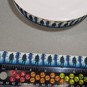 May include: A roll of white satin ribbon with a blue and white print of Bigfoot walking through a forest. The ribbon is 1 inch wide and is measured on a ruler.