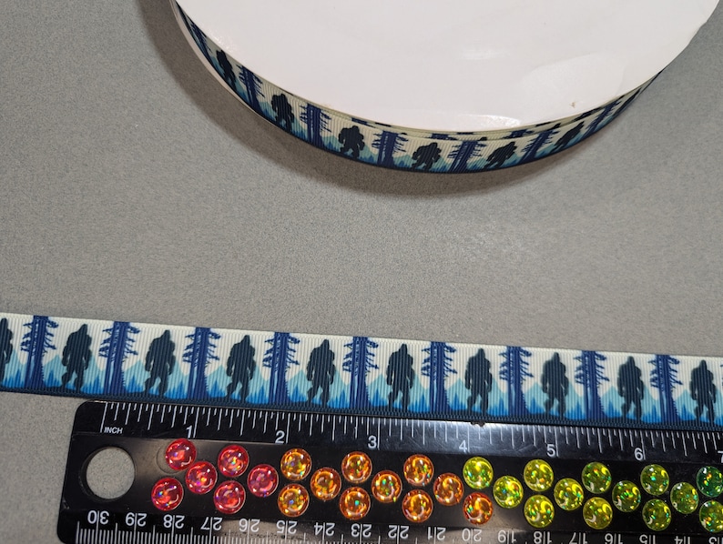 May include: A roll of white satin ribbon with a blue and white print of Bigfoot walking through a forest. The ribbon is 1 inch wide and is measured on a ruler.