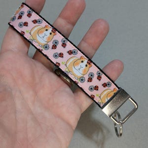 May include: A pink key fob featuring a repeating pattern of guinea pigs, ladybugs, and flowers. The key fob has a metal clasp and key ring. The edges are black.
