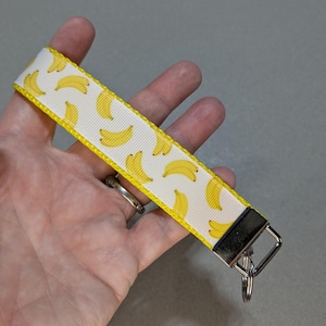 May include: A white keychain wrist strap featuring a pattern of yellow bananas. The strap has a yellow border and a silver-colored metal clasp with a key ring. The strap is held in a hand.