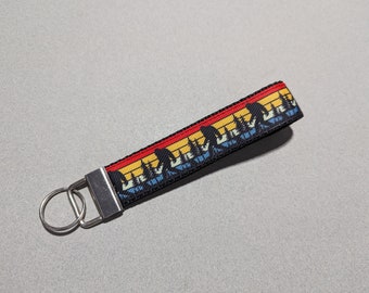 May include: A black key chain with a colorful sunset and silhouette of mountains design. The key chain has a silver metal key ring.