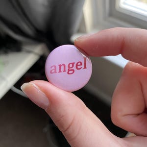 May include: A pale pink button with the word "angel" in a darker pink font. The button is round and held between two fingers. The background includes a window and a grey curtain.