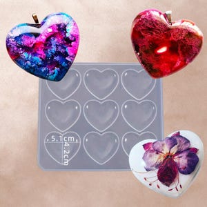 Op de afbeelding: Een transparante siliconen mal met negen hartvormige holtes. De mal is 5,1 cm breed en 4,2 cm hoog. Er zijn drie hars harten met verschillende ontwerpen, waaronder een rood hart met een witte swirl, een paars en blauw hart met een witte swirl en een wit hart met een paarse bloem.