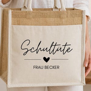 Può includere: Una borsa tote beige con la parte superiore e i manici in iuta naturale. La parte anteriore della borsa ha un pannello bianco con le parole "Schultüte" sopra un cuore e il nome "FRAU BECKER" in corsivo nero.