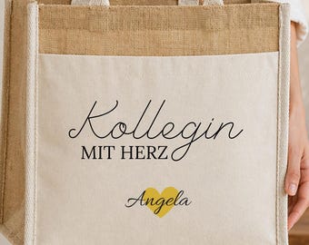 May include: A rectangular tote bag made of natural tan jute and off-white canvas. The front features the black text Kollegin MIT HERZ above a yellow heart icon and the name Angela in black script. It has two sturdy top handles for carrying.