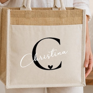 Könnte beinhalten: Eine quadratische Tragetasche aus naturfarbenem Jute- und cremeweißem Canvas-Stoff. Auf der Vorderseite befindet sich ein großer schwarzer Buchstabe C, über den der Name Christina in weißer Schreibschrift gedruckt ist, ergänzt durch ein kleines Herz.