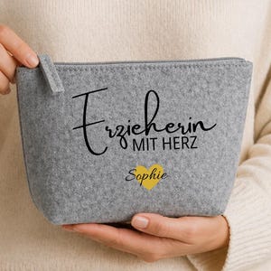 Könnte beinhalten: Eine graue Filz-Reißverschlusstasche mit dem Text "Erzieherin mit Herz" und dem Namen "Sophie" unter einem gelben Herz. Die Tasche wird von einer Person gehalten, die einen cremefarbenen Pullover trägt.
