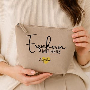 May include: A beige zipped pouch with the text "Erzieherin mit Herz Sophie" in black script and yellow font. The pouch is held by a person wearing a cream-coloured sweater.
