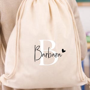 Op de afbeelding: Een beige trekkoordrugzak met de naam "Barbara" in zwart schrift en een zwart hart. De tas heeft witte trekkoorden en wordt over de schouder gedragen. De achtergrond toont een klaslokaal met bureaus en een schoolbord.