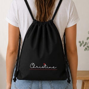 Può includere: Uno zaino nero con chiusura a coulisse che riporta il nome Christine in corsivo bianco con un piccolo cuore rosso sopra la lettera i. La borsa è dotata di spallacci in corda nera ed è indossata sopra una maglietta bianca e jeans blu.