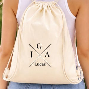 Pode incluir: Uma mochila com cordão de cor creme com texto preto que diz "G", "J", "A" e "Lucas" num design de cruz. A mochila é usada sobre os ombros, com os cordões visíveis. O fundo é verde desfocado.