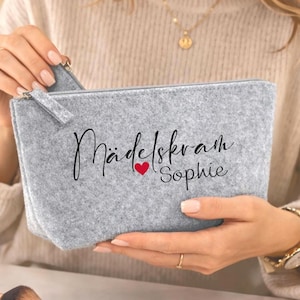 Könnte beinhalten: Eine hellgraue Kosmetiktasche aus Filz mit Reißverschluss. Auf der Tasche steht in schwarzer Schrift "Mädelskram Sophie" mit einem roten Herz. Make-up-Pinsel, ein Spiegel und andere Schönheitsprodukte sind sichtbar.