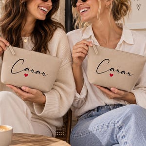 May include: Two beige canvas zip pouches, each featuring the name Canan in black cursive script with a small red heart underneath. The pouches are rectangular with a top zip closure and a neutral, light-coloured fabric texture.