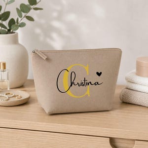 Könnte beinhalten: Eine beige Kosmetiktasche mit Reißverschluss. Die Tasche trägt den Namen "Christina" in Schreibschrift mit einem gelben "C" und einem kleinen schwarzen Herz. Die Tasche steht auf einer Holzoberfläche.