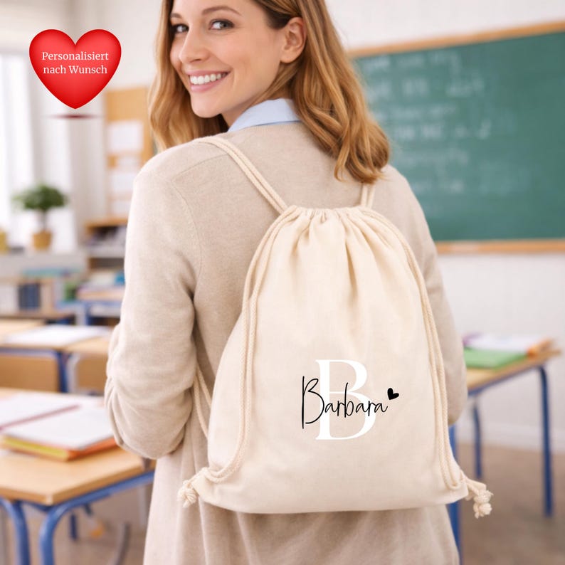 Op de afbeelding: Een beige trekkoordrugzak met de naam "Barbara" in zwart schrift en een zwart hart. De tas heeft witte trekkoorden en wordt over de schouder gedragen. De achtergrond toont een klaslokaal met bureaus en een schoolbord.