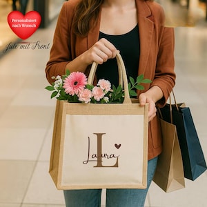 Pode incluir: Uma sacola tote de juta bege com um painel frontal creme. A sacola apresenta um "L" inicial marrom e o nome "Laura" em escrita cursiva, junto com um pequeno coração. A sacola está cheia de flores cor-de-rosa e vegetação.