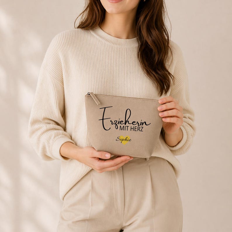 May include: A beige zipped pouch with the text "Erzieherin mit Herz Sophie" in black script and yellow font. The pouch is held by a person wearing a cream-coloured sweater.